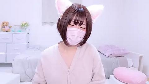 Media: Video of a young Asian woman with short brown hair and cat ears, wearing a pink mask and light gray robe in a white, minimalist bedroom.