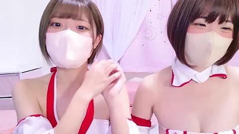 Media: Video of two young Asian women with short brown hair, wearing white surgical masks and red off-shoulder dresses, holding a white lace curtain, set against a pastel-colored background.