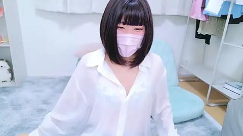 Media: Video of a young East Asian woman with a short black bob, wearing a white button-up shirt, face mask, and sitting on a light blue bean bag chair in a white room with shelves and clothes.