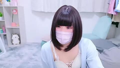 Media: Video of a young East Asian woman with straight black hair and blunt bangs, wearing a white face mask, light blue cardigan, and a cream lace bra, sitting in a pastel-colored room with a plush gray rug.