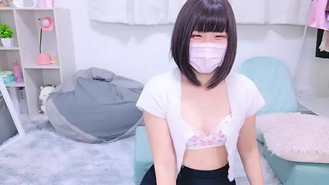 Media: Video of an East Asian woman with straight black hair, wearing a white mask, white top, and black pants, sitting on a gray beanbag in a pastel-colored room.