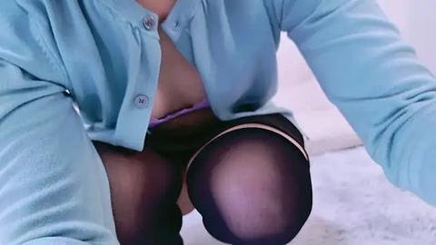 Media: Video of a woman with light skin, wearing a blue button-up shirt unbuttoned, revealing a purple bra, black sheer stockings, and sitting on a white bed.