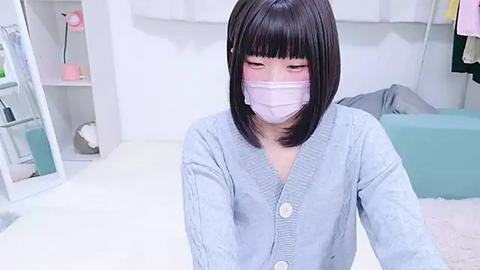 Media: Video of an Asian woman with straight, black hair, wearing a light blue cardigan and a pink face mask, sitting in a minimalist room with white walls, a shelf, and a teal couch.