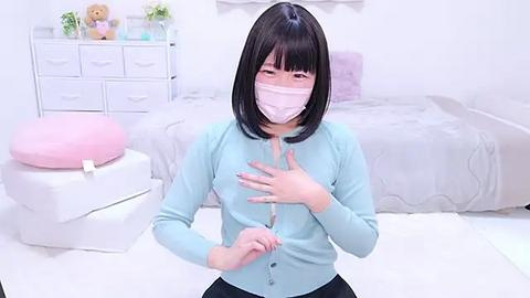 Media: Video of an East Asian woman with straight black hair, wearing a light blue cardigan, face mask, and a pink shirt, standing in a minimalist, white bedroom with a bed, dresser, and stuffed toys.