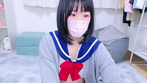 Media: A video of an Asian woman with straight black hair, wearing a grey sailor school uniform with a red bow, white mask, and grey cardigan, kneeling on a grey carpeted floor.