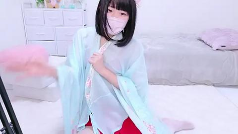 Media: Video of an Asian woman with straight black hair, wearing a light blue kimono with pink floral patterns, a white mask, and red pants, standing in a minimalist, white-walled bedroom with a bed and dresser.