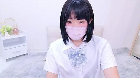 Media: Video of a young Asian woman with short black hair, wearing a white blouse, plaid bow, and pink face mask, sitting on a gray shag rug in a minimalist, white room.