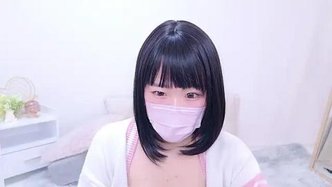 Media: Video of an East Asian woman with straight black hair and bangs, wearing a white face mask, pink cardigan, and a white top, standing in a minimalist room with a white wall, a shelf with potted plants, and a grey rug.