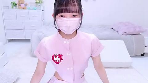 Media: A video of an East Asian woman with pale skin and straight black hair, wearing a light pink nurse outfit with a red heart patch, standing in a minimalist, white bedroom with a bed and dresser.