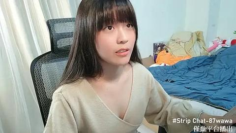 Media: Video of a young Asian woman with straight, shoulder-length dark hair, wearing a beige V-neck sweater, sitting on a black office chair in a cluttered bedroom with unmade bed and pink teddy bear.