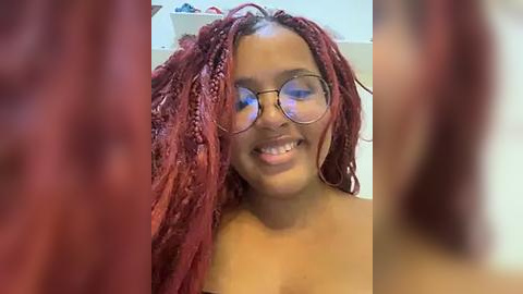 Media: Video of a smiling Black woman with medium-dark skin tone and long, thick, maroon dreadlocks. She wears large round glasses and is topless, revealing her bare chest. Background shows a blurred white room with shelves.