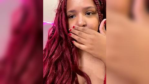 Media: Video of a young Black woman with long, wavy, maroon braids, partially covering her mouth with one hand. She has light brown skin and red nail polish.