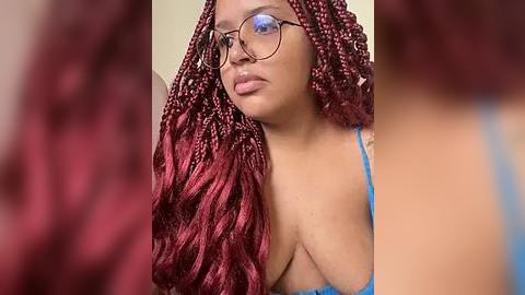 Media: Video of a Black woman with medium skin tone, wearing round glasses, blue bra, and voluminous, deep red braids cascading over her shoulders. Background is blurred.