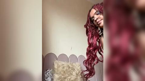 Media: A video of a young woman with long, wavy, red hair, wearing black-framed glasses, standing in a cozy, cream-colored room with a white, fluffy pillow.