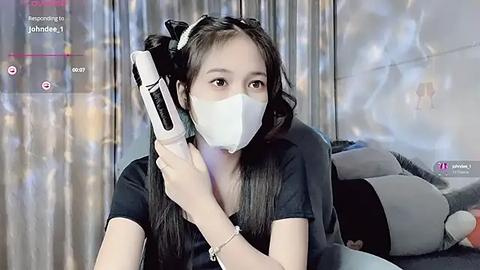 Media: A video of an Asian woman with long black hair, wearing a white mask, holding a white phone to her ear, in a modern bedroom with gray curtains and plush pillows.