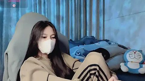 Media: Video of a young woman with long black hair, wearing a white mask, beige jacket, and black pants, sitting on a blue couch with plush toys in a room with light blue curtains.