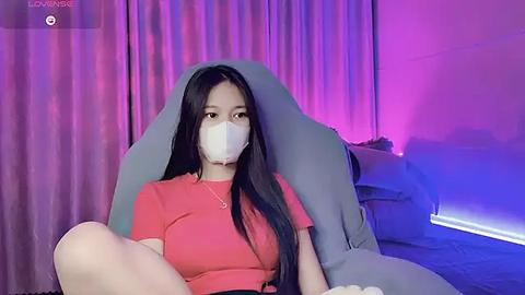 Media: Video of an Asian woman with long black hair, wearing a white mask and a red T-shirt, sitting on a grey gaming chair in a dimly lit room with purple and blue lighting.