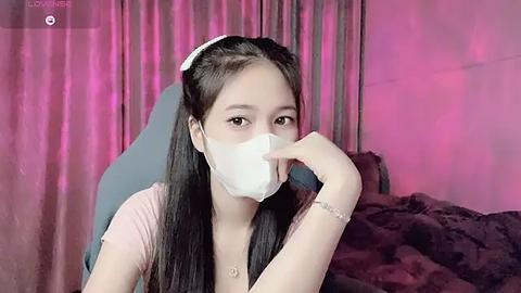 Media: Video of an Asian woman with long black hair, wearing a pink top, white mask, and headband, sitting on a chair against pink curtains and dark bedding.