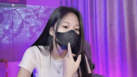 Media: Video of an Asian woman with long black hair, wearing a black face mask and a white t-shirt, in a room with purple and white curtains, miming a phone call.