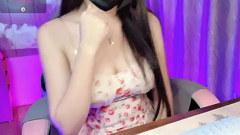 Media: Video of a young Asian woman with long dark hair, wearing a strapless floral dress, seated at a table, with a black mask covering her face, under purple lighting.