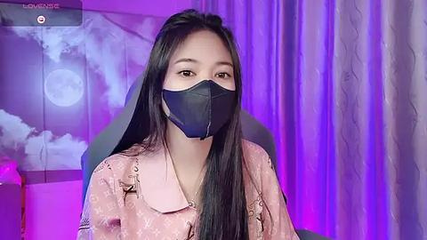 Media: Video of an Asian woman with long black hair, wearing a pink floral blouse and black face mask, seated indoors with purple and blue lighting, background featuring moon and cloud wall art.