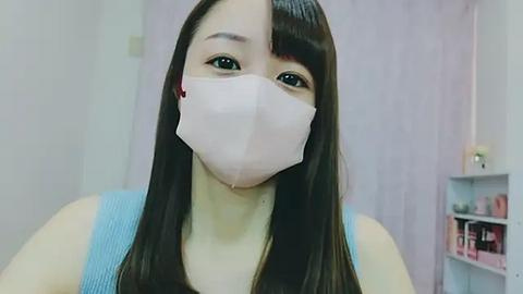 Media: A video of an East Asian woman with long black hair, wearing a white face mask, light blue tank top, and standing in a modern, softly lit room with a pink wall and white shelving unit.