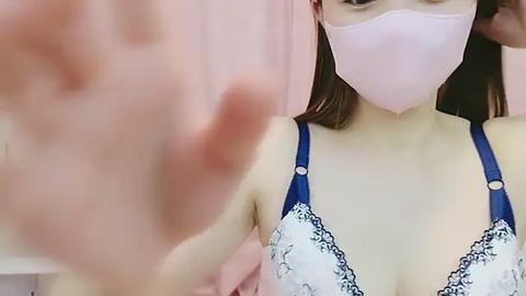 Media: Video of an Asian woman wearing a light pink face mask and a blue and white lace bra, with her hand partially in frame, indoors with pink curtains.