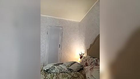 Media: Video of a dimly lit bedroom with light beige walls, a floral-patterned bedspread, and a small nightstand lamp. The room has a white door and a partially visible headboard.