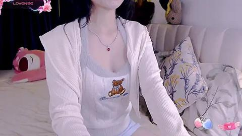 Media: A video of a young woman with pale skin, wearing a white ribbed cardigan over a white tank top with a cartoon bear graphic. She has long black hair and is seated on a bed with floral-patterned pillows.