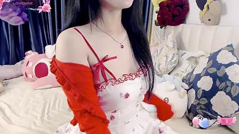 Media: A video of an Asian woman with long black hair, wearing a red bow-tie top, red cardigan, and necklace, sitting on a bed with plush toys.