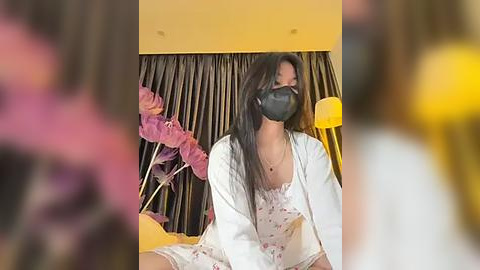 Media: Video of an Asian woman with long black hair, wearing a floral dress, mask, and robe, seated on a bed with pink flowers and a lamp in the background.