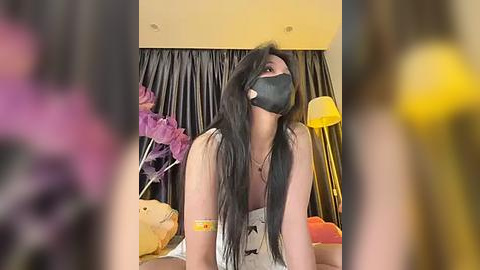 Media: Video of a woman with long black hair and fair skin wearing a black face mask, sitting on a bed with a beige headboard, surrounded by purple and orange pillows, and a yellow lamp.