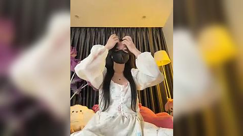 Media: Video of an Asian woman with long black hair, wearing a white dress, black mask, and glasses, adjusting her hair, in a room with dark curtains, plush toys, and a lamp.