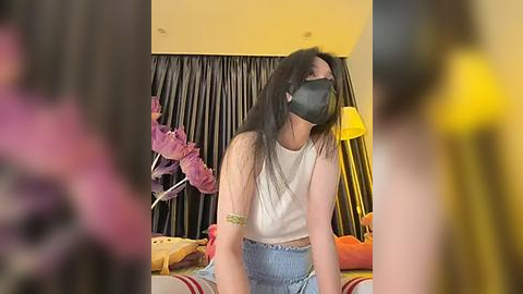 Media: Video of a young woman with long black hair, wearing a black face mask, white tank top, and blue shorts, sitting on a bed with yellow pillows, in a dimly lit room with dark curtains.