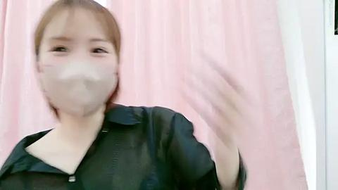 Media: A blurred video of an Asian woman with light skin and short brown hair, wearing a black leather jacket and a white mask, waving her hand against a pink curtain background.