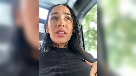 Media: Video of a Latina woman with long, straight black hair, wearing a black ribbed top, sitting in a car, with blurred foliage and a blurred person in the foreground.