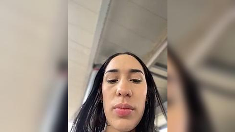 Media: Video of a young woman with long, straight black hair and light brown skin, wearing a neutral expression, taken from a low angle. Background shows a blurred indoor ceiling and pipes.