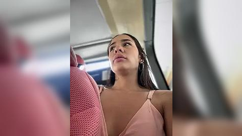 Media: Video of a young Latina woman with straight dark hair, light skin, and a pink dress, looking up in a bus, with blurred pink seats and a window in the background.