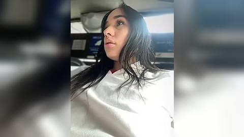 Media: Video of a woman with long, wet, black hair, wearing a white robe, sitting in a car, blurry background, looking thoughtful.