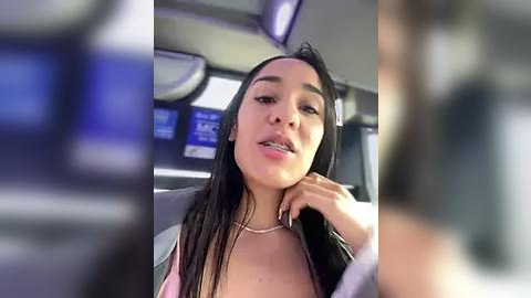 Media: Video of a Latina woman with medium skin tone, long black hair, and silver necklace, seated in a car, wearing a pink top, looking relaxed.