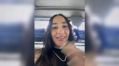 Media: Video of a smiling Latina woman with long dark hair, wearing a black sleeveless top, inside a car, with blurred background.