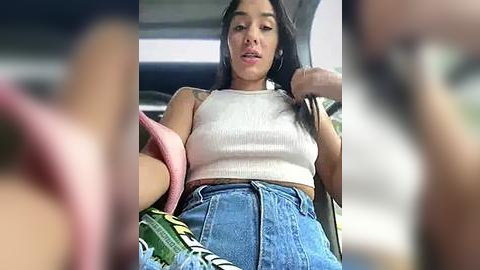 Media: Video of a young woman with long black hair, wearing a white ribbed crop top and high-waisted blue jeans, sitting in a car, her hand in her hair, blurry background.