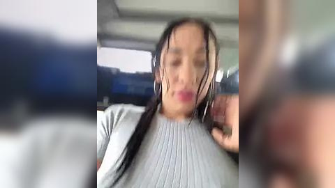 Media: A blurred video of a young woman with wet hair, wearing a grey sweater, sitting in a car with a blurred background.