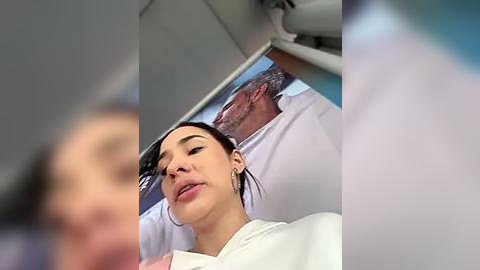 Media: Video of a smiling woman with dark hair and light skin, wearing a white top, seated next to an older man with gray hair and beard, both in a vehicle.