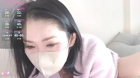 Media: Video of an East Asian woman with straight black hair, wearing a white face mask and light pink robe, lying on a bed with gray sheets, in a softly lit room.