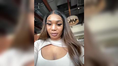 Media: Video of a Black woman with long, straight, ombre hair, wearing a beige, crop-top dress, with a low-cut neckline revealing cleavage, standing indoors with a blurred background featuring exposed ceiling beams and a chandelier.