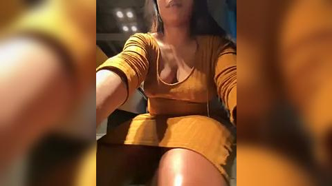 Media: Video of a woman with light brown skin, wearing a tight mustard yellow dress, sitting in a dimly-lit room with blurred background.