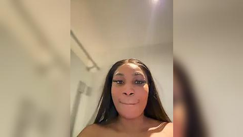 Media: A video of a smiling Black woman with long, straight black hair, wearing heavy eye makeup, standing indoors in a bathroom with white walls and a metal towel rack.