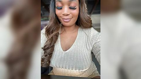 Media: Video of a smiling, dark-skinned Black woman with long, wavy brown hair, wearing a beige V-neck sweater. Background is blurred, focusing on her serene expression and casual outfit.