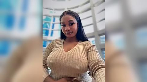 Media: Video of a young Black woman with straight, long black hair, wearing a beige ribbed sweater, standing in a modern, bright interior with curved walls and blue accents.